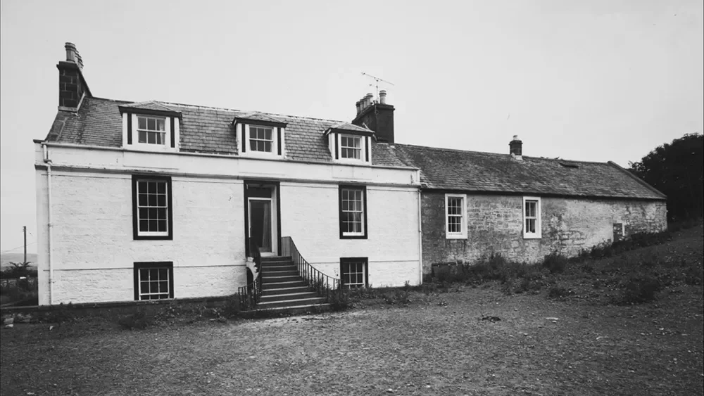 Black and white photo of a two-story house with a staircase leading up to the front door, surrounded by an unpaved yard.
