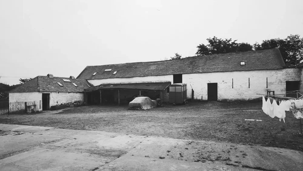 A black and white photo of a rustic farm building with a large open yard, laundry hanging on a clothesline, and trees in the background.