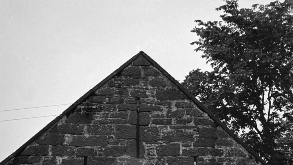 The top of a brick house with a gable roof, a large tree in the background, and a partly cloudy sky.