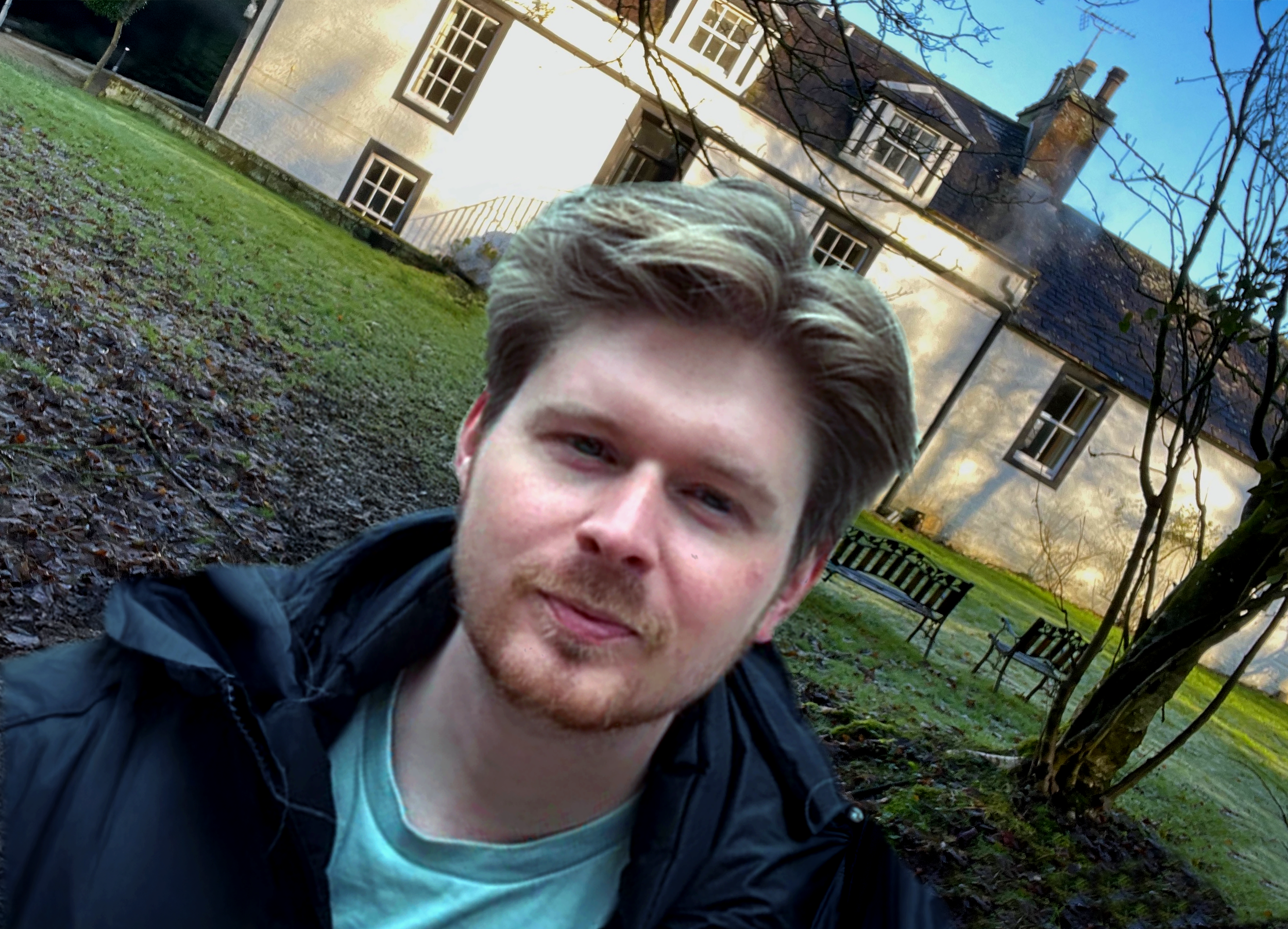 A young man wearing a dark jacket, slightly smiling, standing in front of an old house