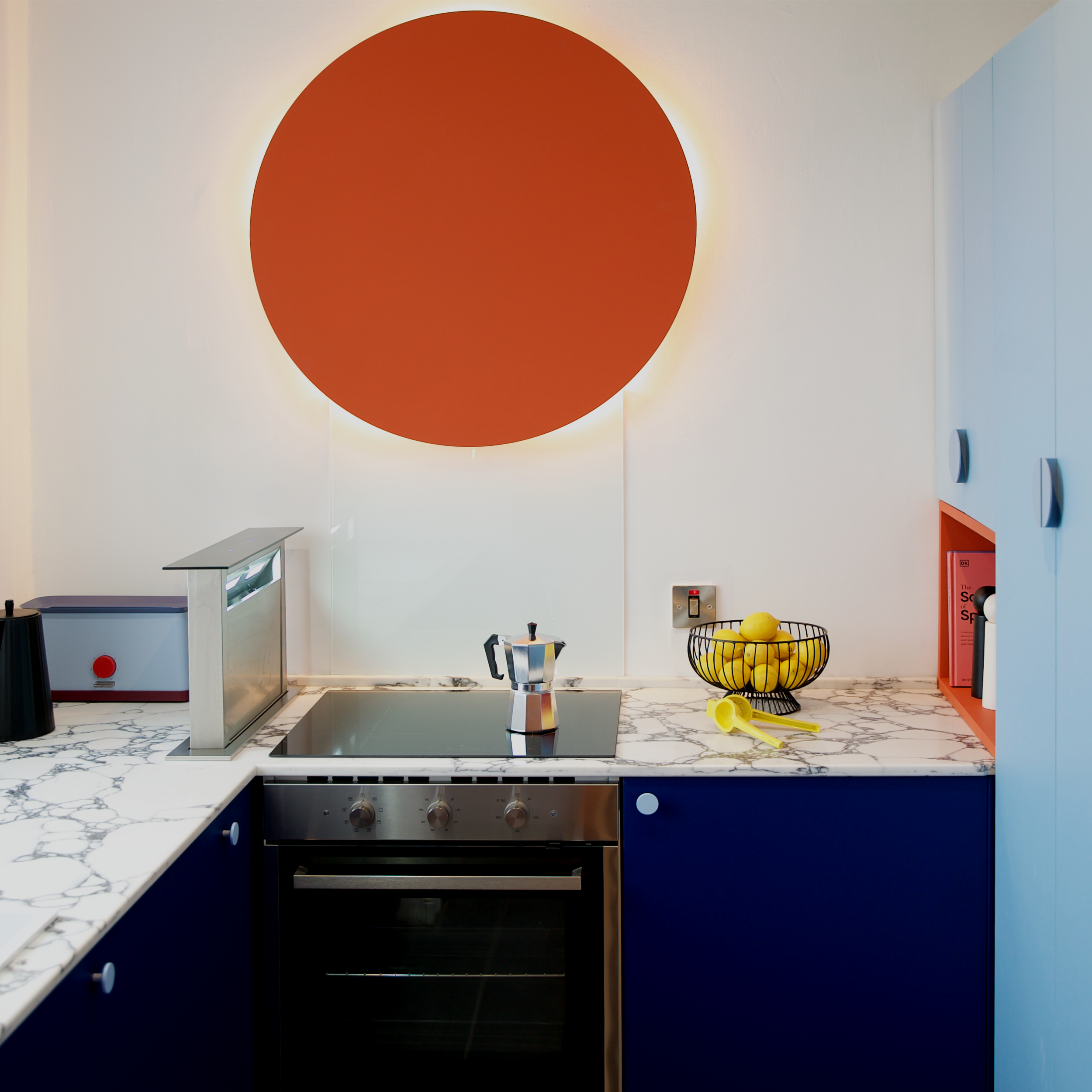 A modern kitchen with a stovetop, a coffee pot, a basket of lemons, and colorful cabinets. A large orange circular lamp with backlighting is on the wall.