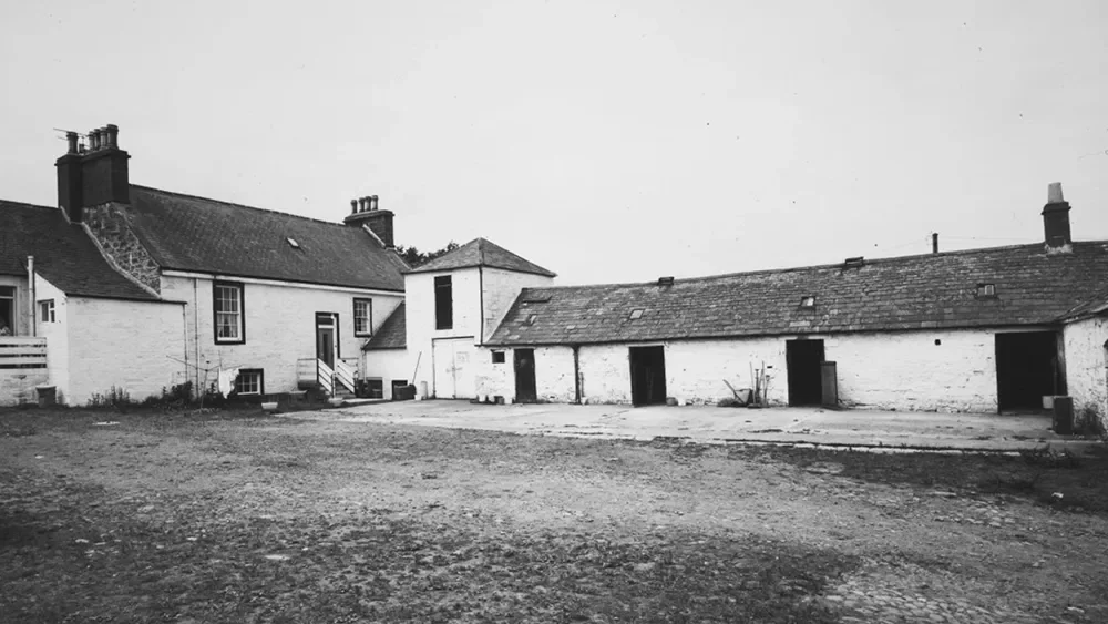 Black and white photo of a rustic farmyard with old buildings, some with tiled roofs, and open doorways, surrounded by a dirt ground.