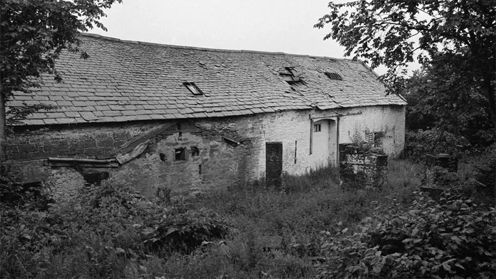 An old, abandoned stone building with a sloped roof, surrounded by overgrown vegetation and trees.