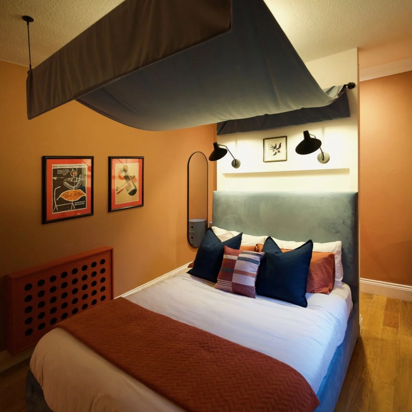 A cozy bedroom with a bed decorated with blue and patterned pillows, a blue headboard, and warm lighting. The wall behind has framed artwork, and a canopy hangs from the ceiling above the bed.