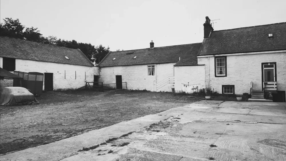 Black and white photo of a rural courtyard with old white houses, a paved area, a car covered with a tarp, and a small greenhouse.