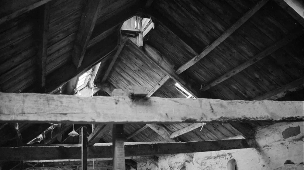 Black and white photo of an attic or loft with exposed wooden beams and a slanted ceiling, some light coming through gaps in the roof.