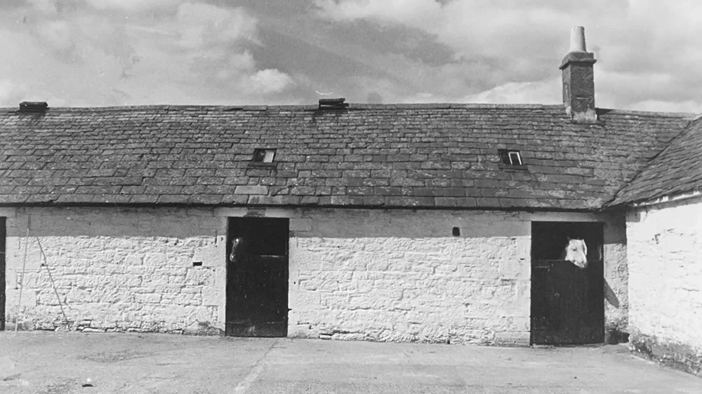 Black and white photo of a rustic brick building with a sloped roof, two small skylights, and a chimney, with a person peering out from behind a door on the right side.