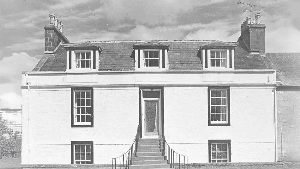 A black and white photo of a two-story house with a stairway leading to the front door. The house has a pitched roof and three dormer windows on the upper level. There are four large windows on the main level, two on each side of the front door, and two smaller basement windows.