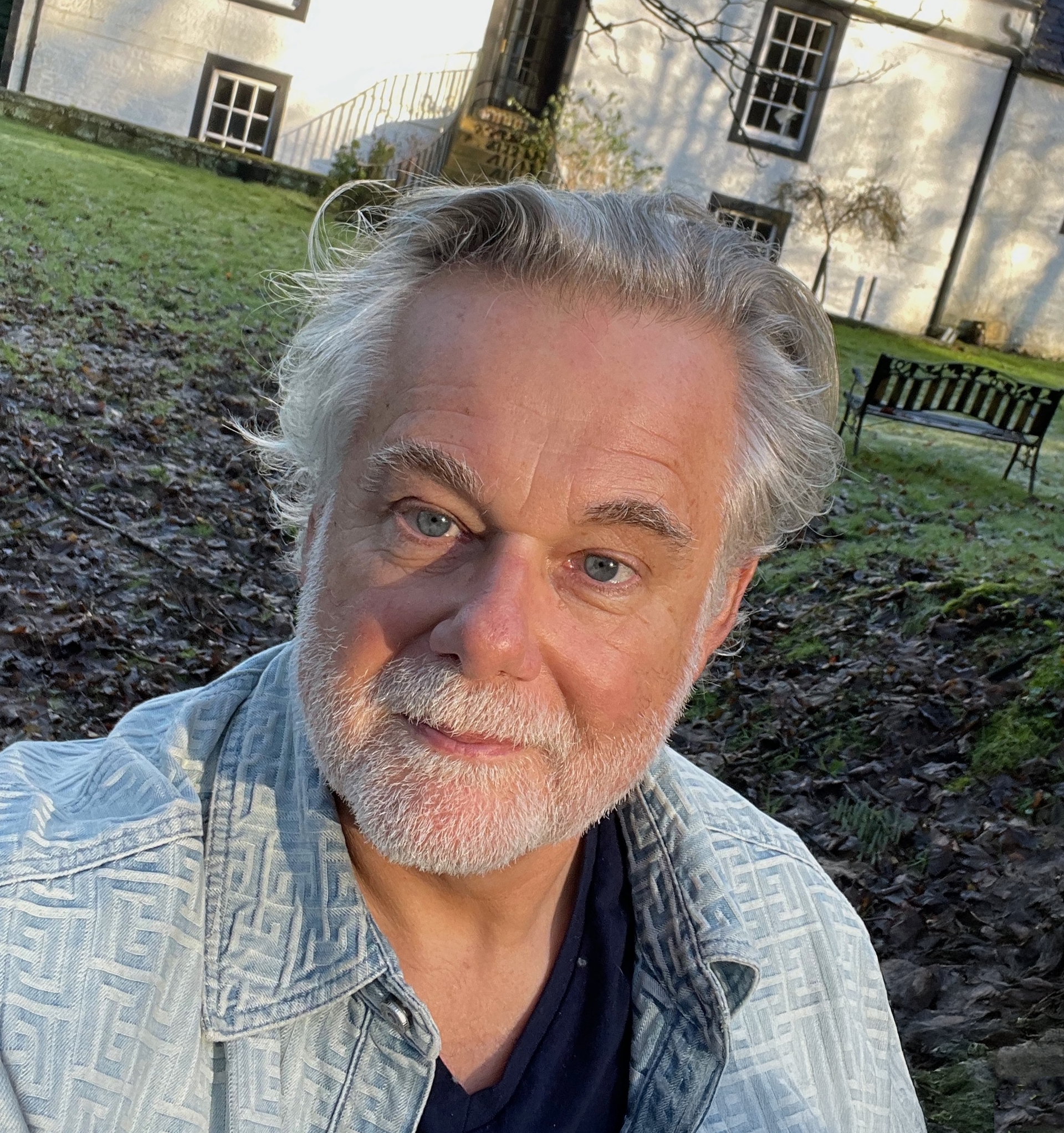 Close-up of an elderly man with gray hair and blue eyes, wearing a blue patterned shirt, outdoors in front of a house with a small front porch, black window frames, a white wall, and leafless trees, with benches and a lawn in the background.