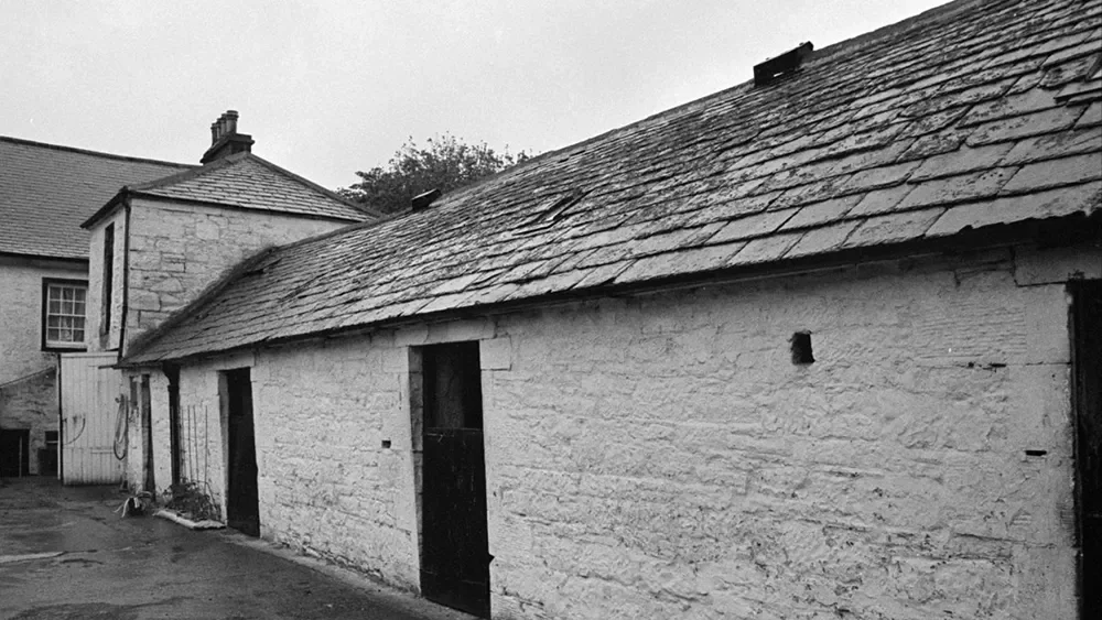 Black and white photo of an old stone building with a sloped roof, small windows, and at least one door.