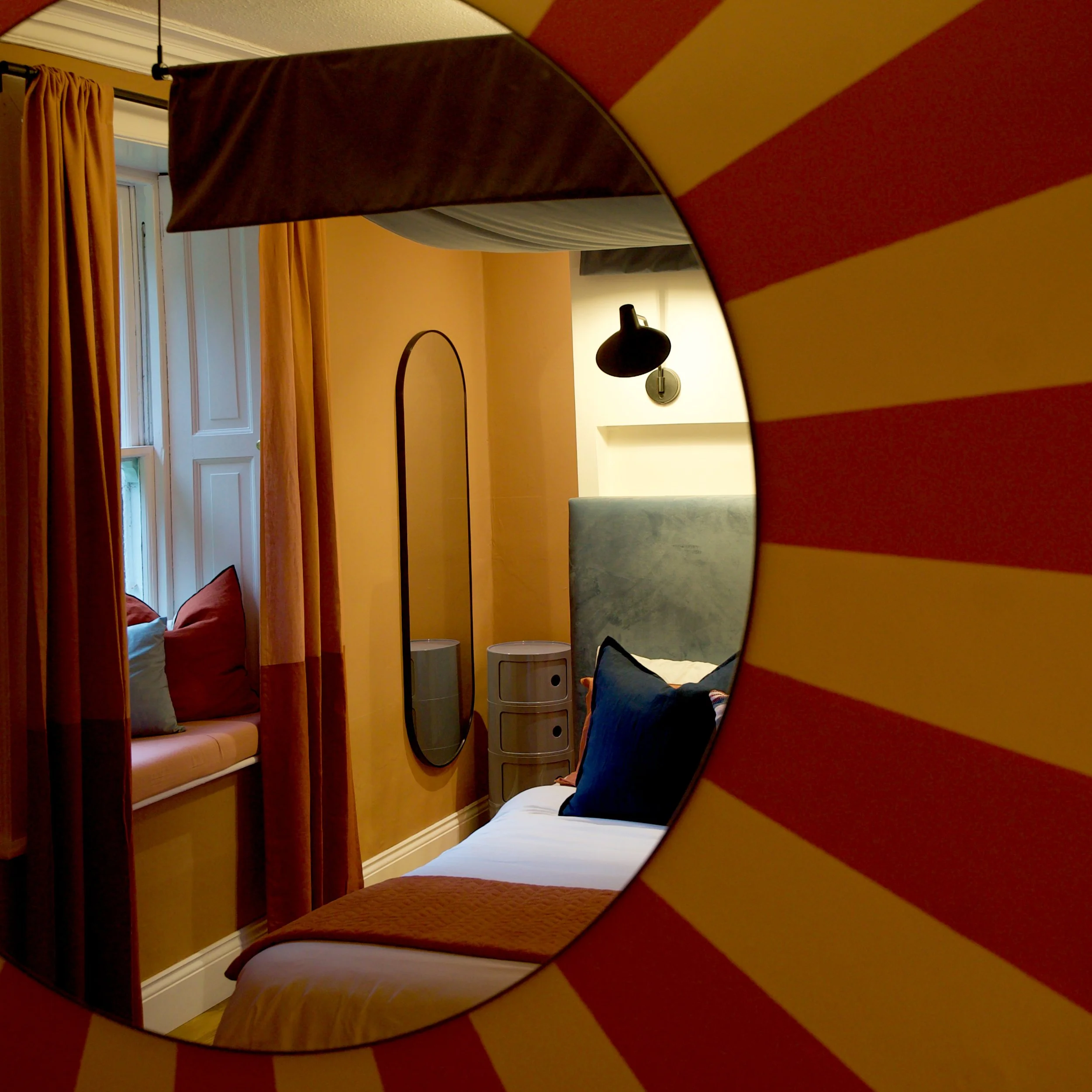 Close-up of a mirror reflecting a corner of a cozy room with a bed, yellow walls, and a black wall lamp.