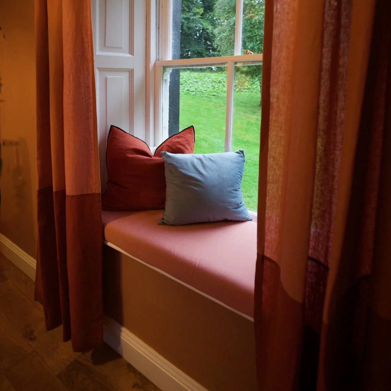A cozy window seat with two pillows, one red and one light blue, in front of a large window overlooking a green outdoor area.