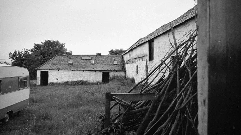 A black and white photo of a rural scene with old buildings, a caravan on the left, and a pile of stick or branches on the right.