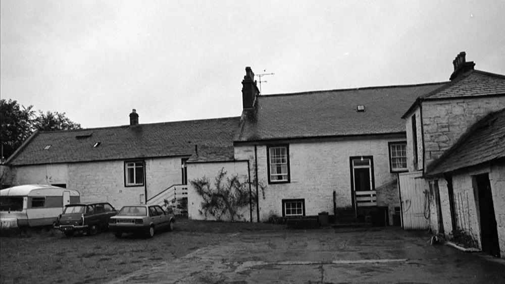 Black and white photo of a courtyard with three parked cars, a trailer, and a stone building with multiple windows and chimneys.