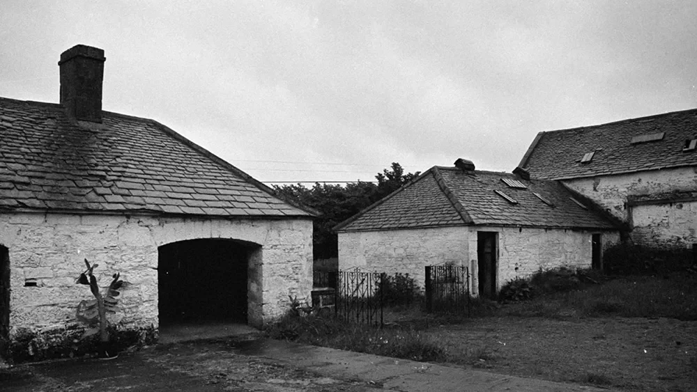 Black and white photo of two old stone buildings with tiled roofs, one with an arched opening and the other with small skylights, surrounded by a yard and a metal fence.