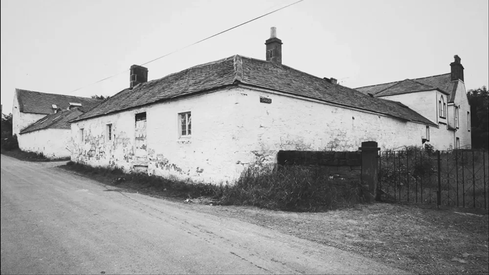 Black and white photo of an old, worn-out white cottage with a sorry-looking front door and small windows. The roof is pitched with a chimney, and the house is situated along a dirt road with overgrown grass around it.
