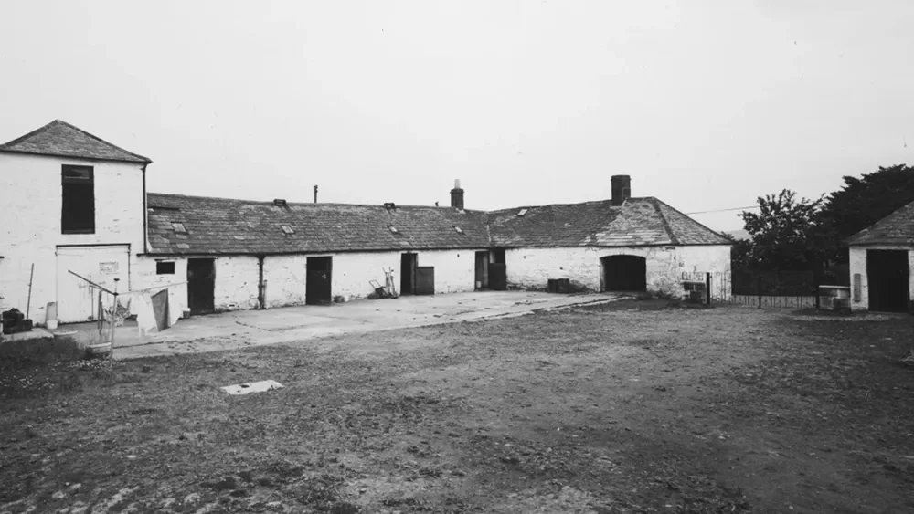 Black and white photo showing an old farmyard with several rustic barns and a large open dirt area.