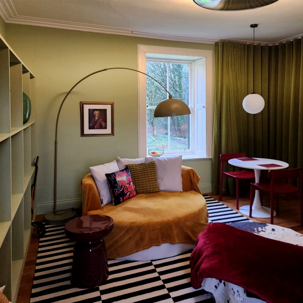Cozy living room with a mustard colour sofa, striped black and white rug, green walls, and a window with green curtains. There is a small round side table, arched floor lamp, framed painting, and a small dining table with red chairs.