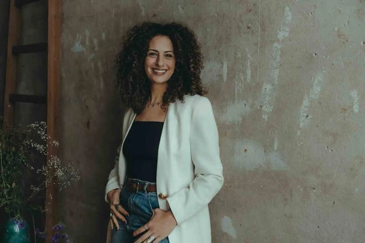 Jasmin, lachend mit lockigem Haar, trägt eine weiße Jacke und ein schwarzes Top, steht vor einer verputzten Wand.