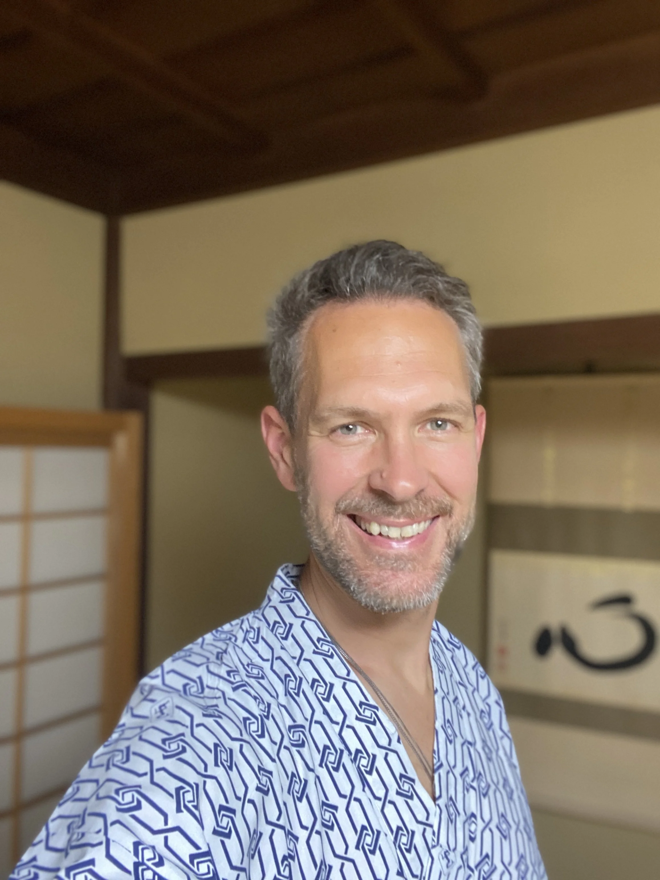 Un homme souriant avec des cheveux gris, portant une chemise à motifs géométriques bleus et blancs, dans une pièce avec des murs en bois et un panneau japonais en arrière-plan.