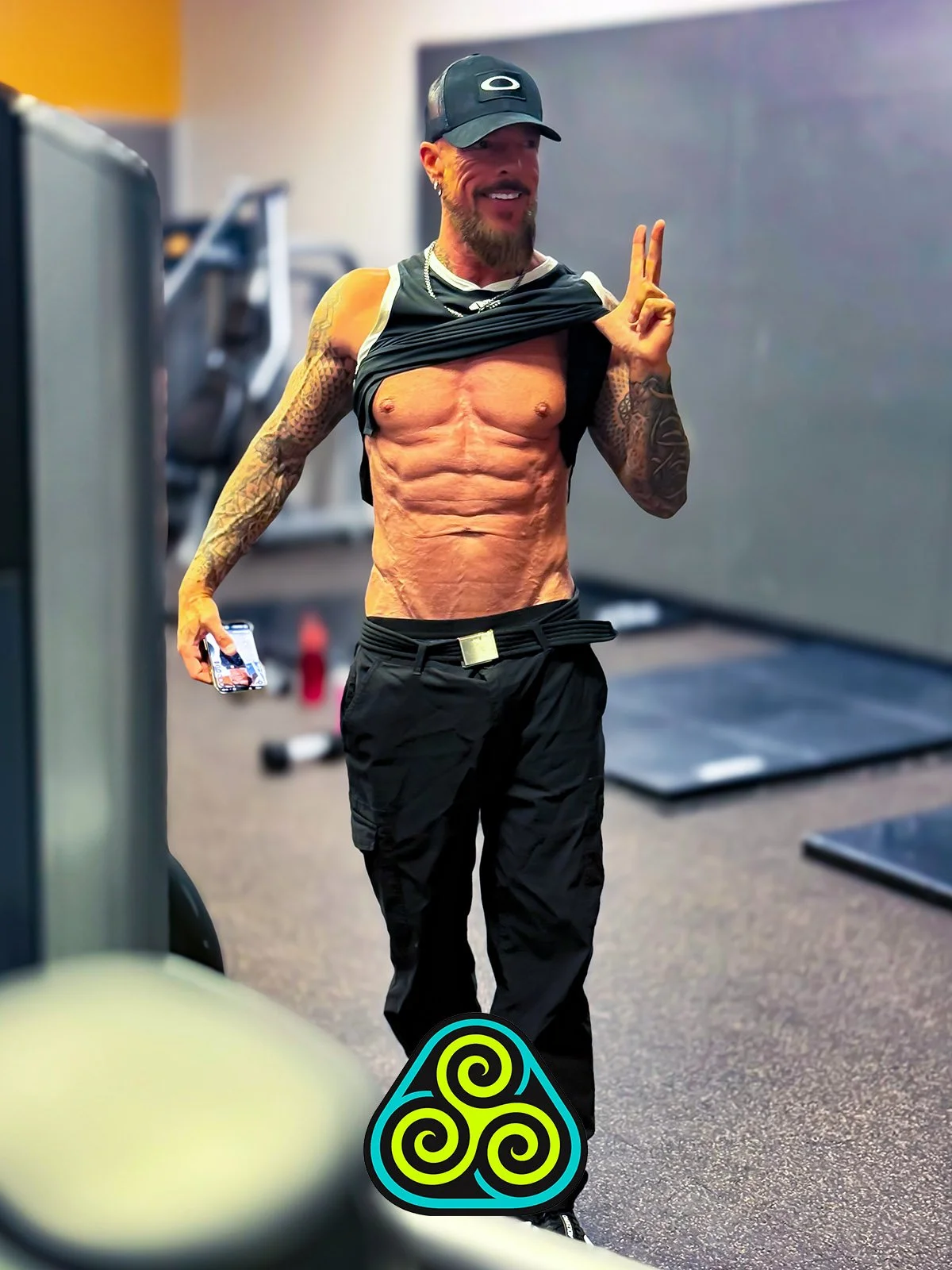A shirtless man with tattoos on his arms and a beard, wearing a black cap and black pants, holding a phone in one hand and making a peace sign with the other, in a gym.