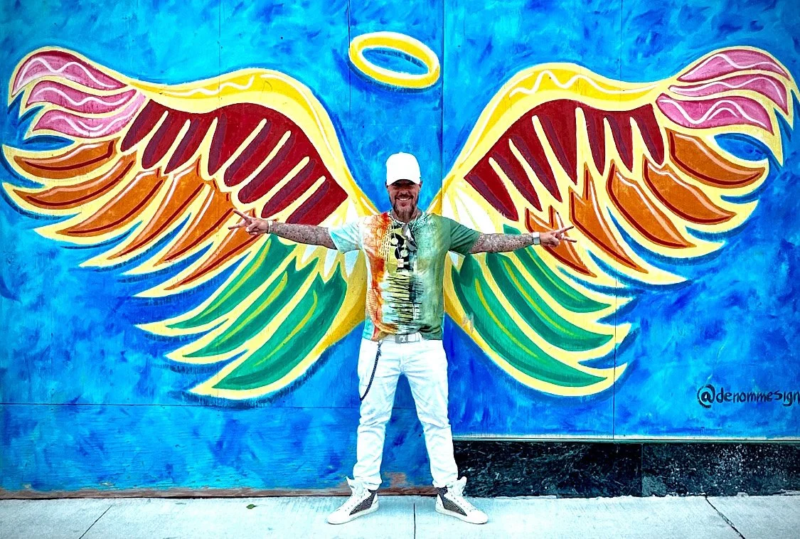 James Erdt standing with arms outstretched in front of a colorful mural of angel wings painted on a wall.