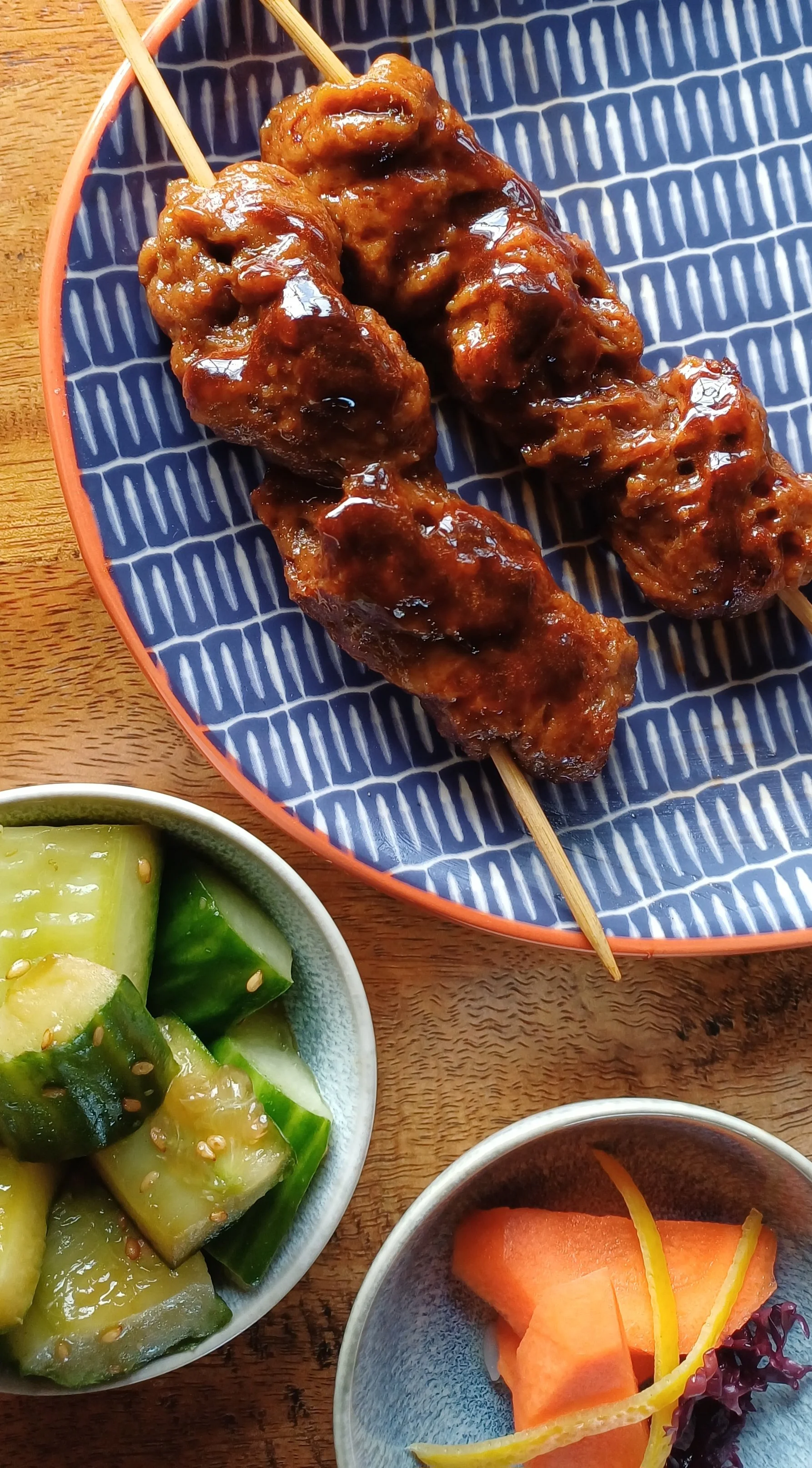 Plant-based yakitori-style seitan skewers with glossy glaze, served with cucumber salad and Japanese pickles on a blue plate
