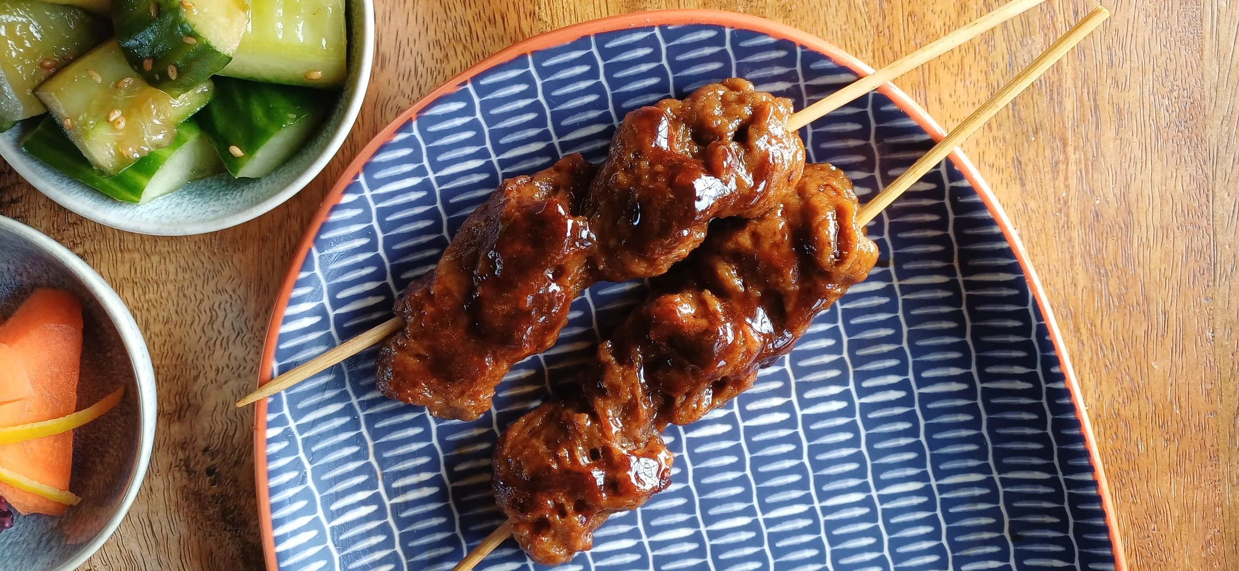Close up of plant-based yakitori-style seitan skewers with glossy glaze, served with cucumber salad and Japanese pickles on a blue plate