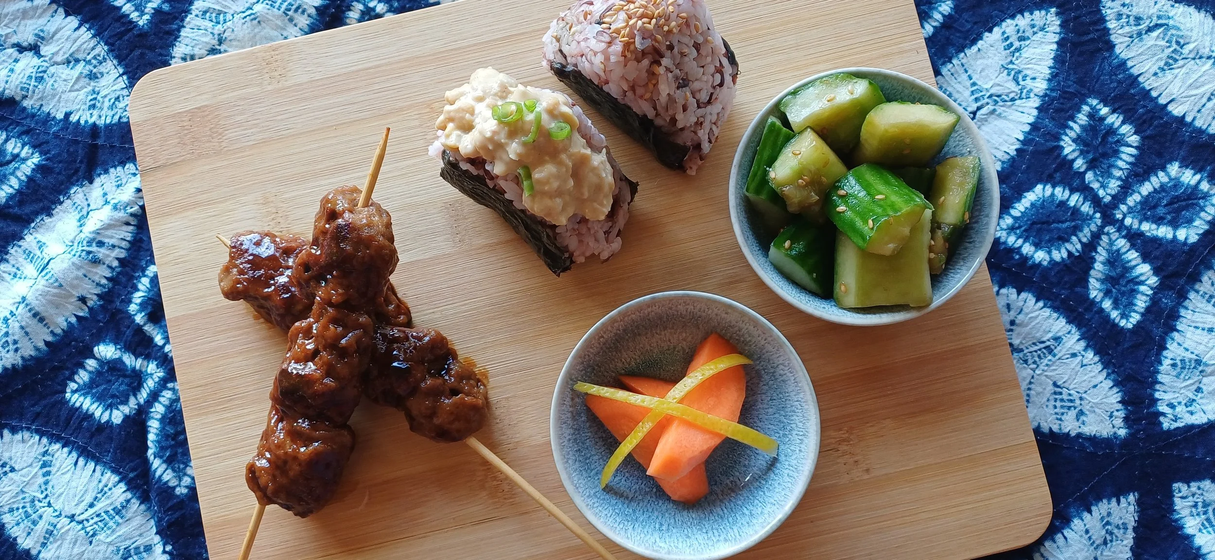 Japanese plant-based bento with onigiri, glazed seitan skewers and small bowls of fresh vegetables and pickles