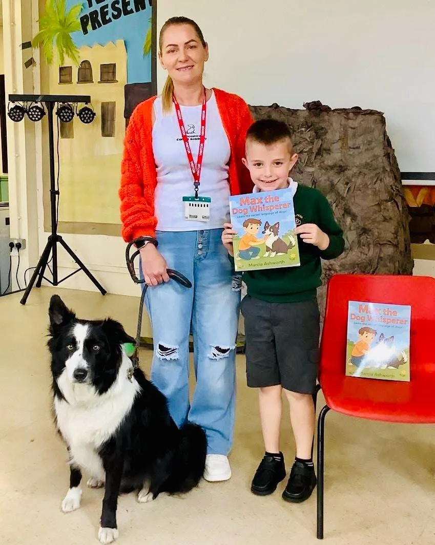 A woman and a boy holding a children's book titled 'Max the Dog Whisperer' stand indoors with a border collie dog sitting on the floor. The woman is wearing a red cardigan and ripped jeans, and the boy is dressed in a green sweater and shorts. There is a red chair with another copy of the book on it, and a banner in the background with part of the word "PRESENT" visible.