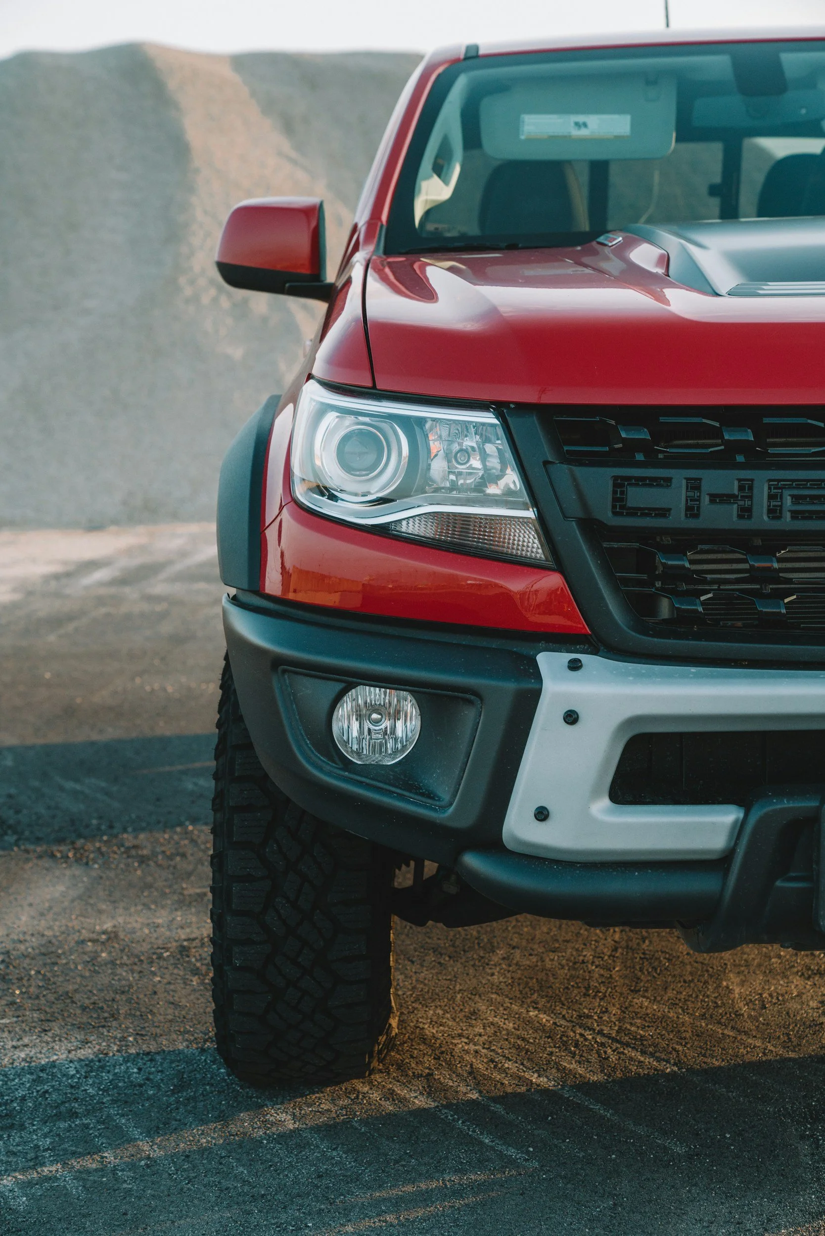 Close-up of the front part of a red pickup truck with a rugged tire, parked on a dirt road with a mountain landscape in the background.