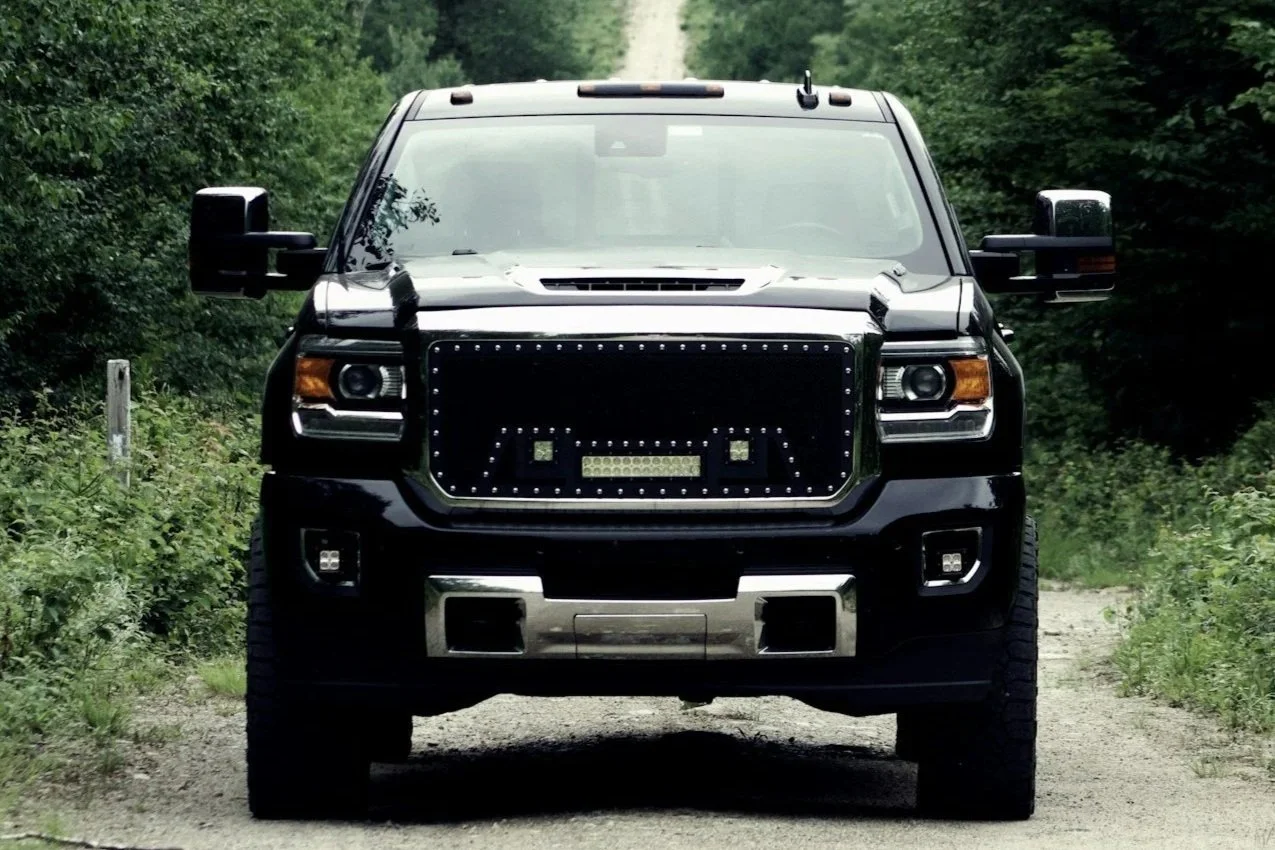 A black pickup truck with a custom LED light grille driving on a dirt path surrounded by green trees.