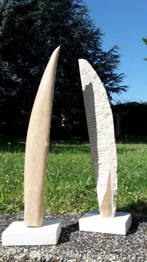 Zwei abstrakte Skulpturen aus Stein stehen auf einem Gehweg, vor einem grünen Rasen und Bäumen, unter blauem Himmel.
