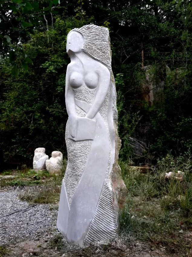 Skulptur einer nackten Frau mit langen Haaren, die eine Tasche hält, vor einem bewaldeten Hintergrund.