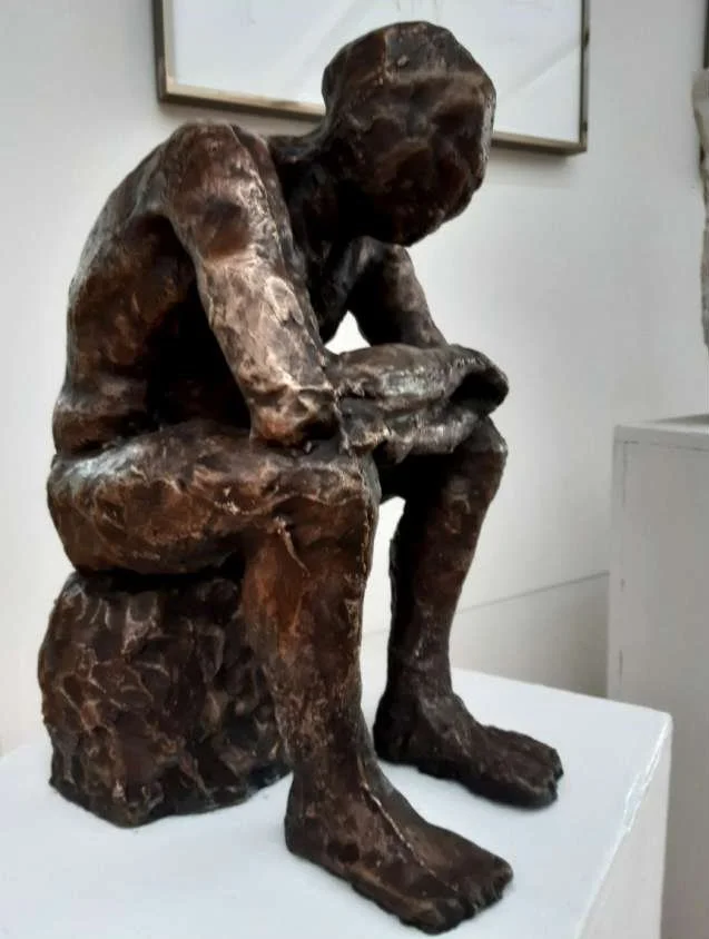 Bronzefigur eines sitzenden Jungen mit hockender Haltung auf einer Plattform.