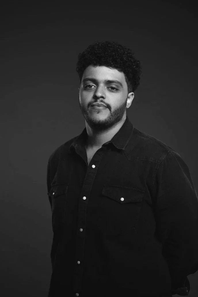 Black and white portrait of a young man with curly hair, beard, wearing a button-up shirt, standing against a plain dark background.