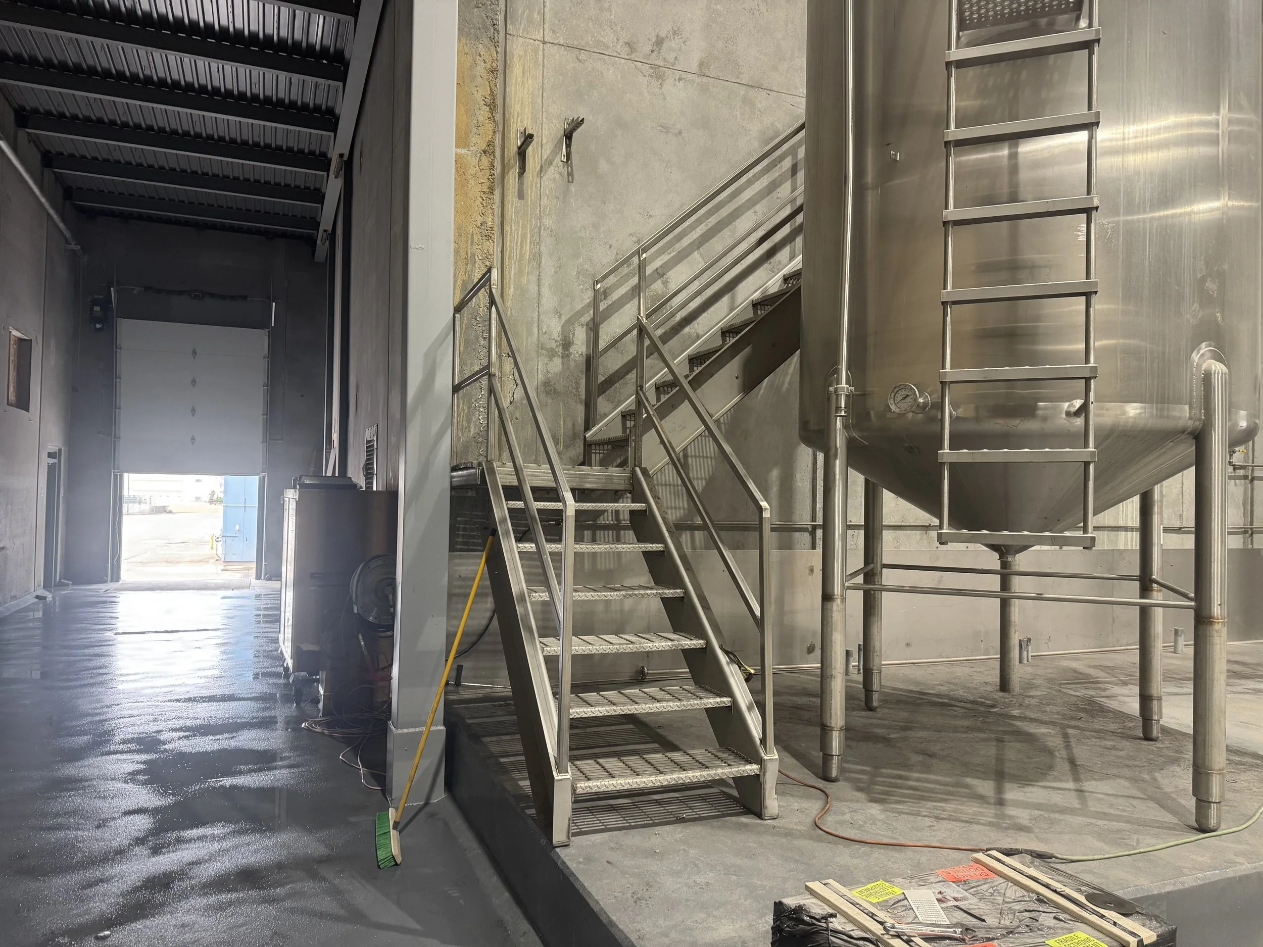 Industrial interior with metal stairs, stainless steel tank, and various tools on the floor. Bright doorway at the end of the room.