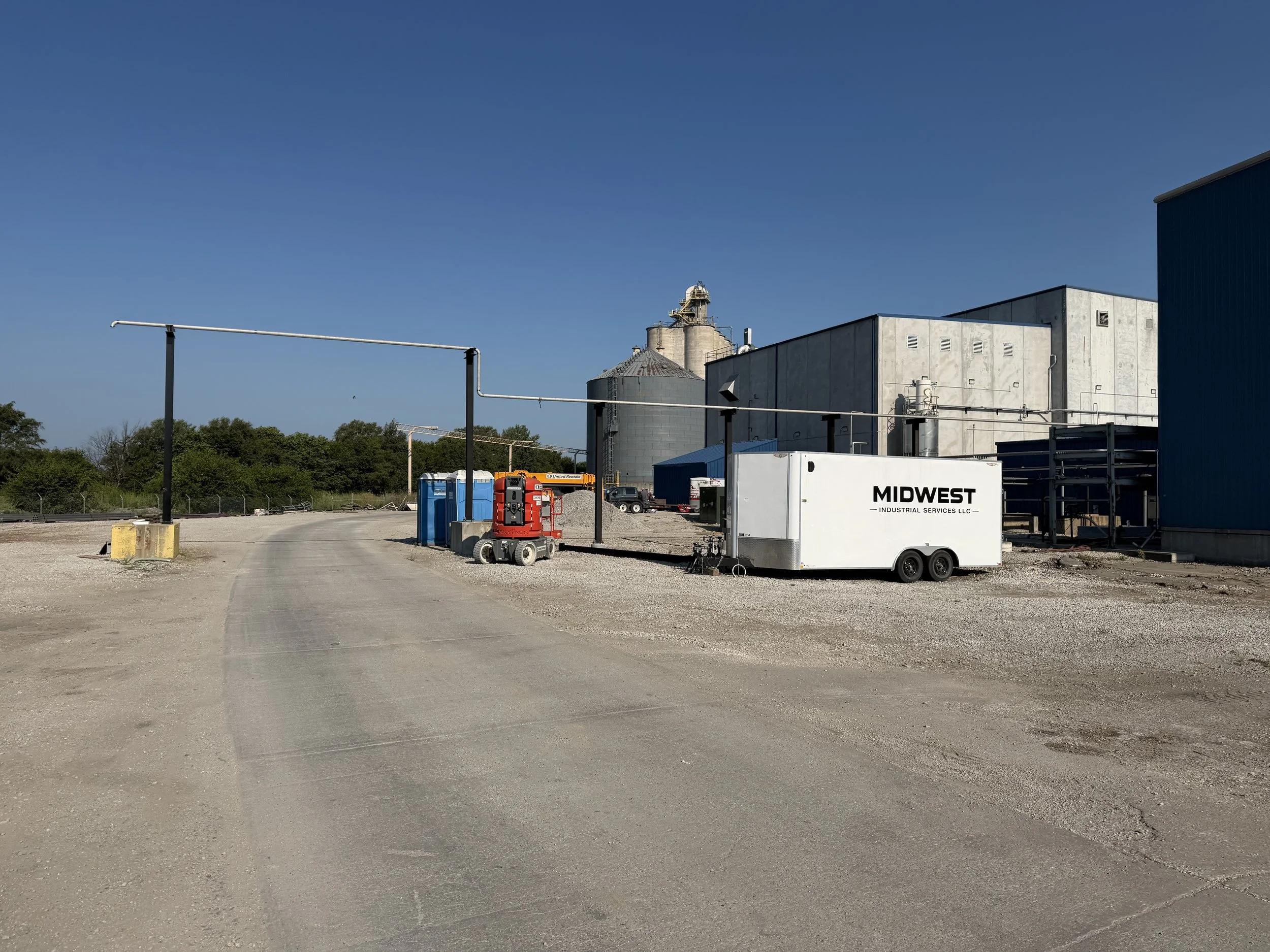 Industrial area with storage tanks, equipment, and a white trailer labeled 'MIDWEST INDUSTRIAL SERVICES LLC'; gravel ground and trees in the background under a clear blue sky.