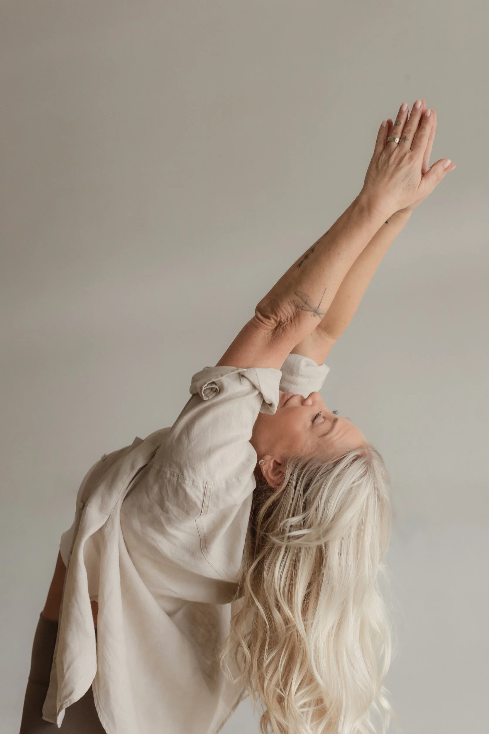 A woman with long, wavy blonde hair lying on her back, wearing a loose, light-colored shirt with rolled-up sleeves, reaching up with both hands towards a plain, light background.