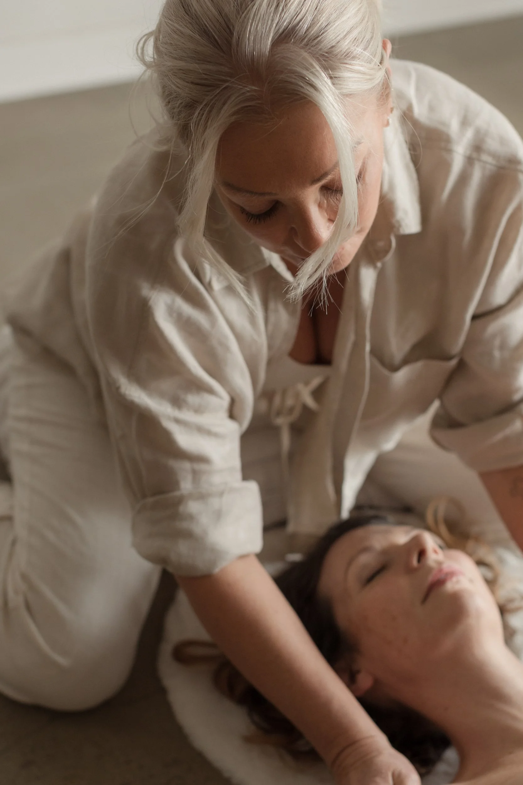 Two women in light-colored clothing lying on the floor, with one woman resting her head on the other's stomach, eyes closed, creating a peaceful and intimate scene.