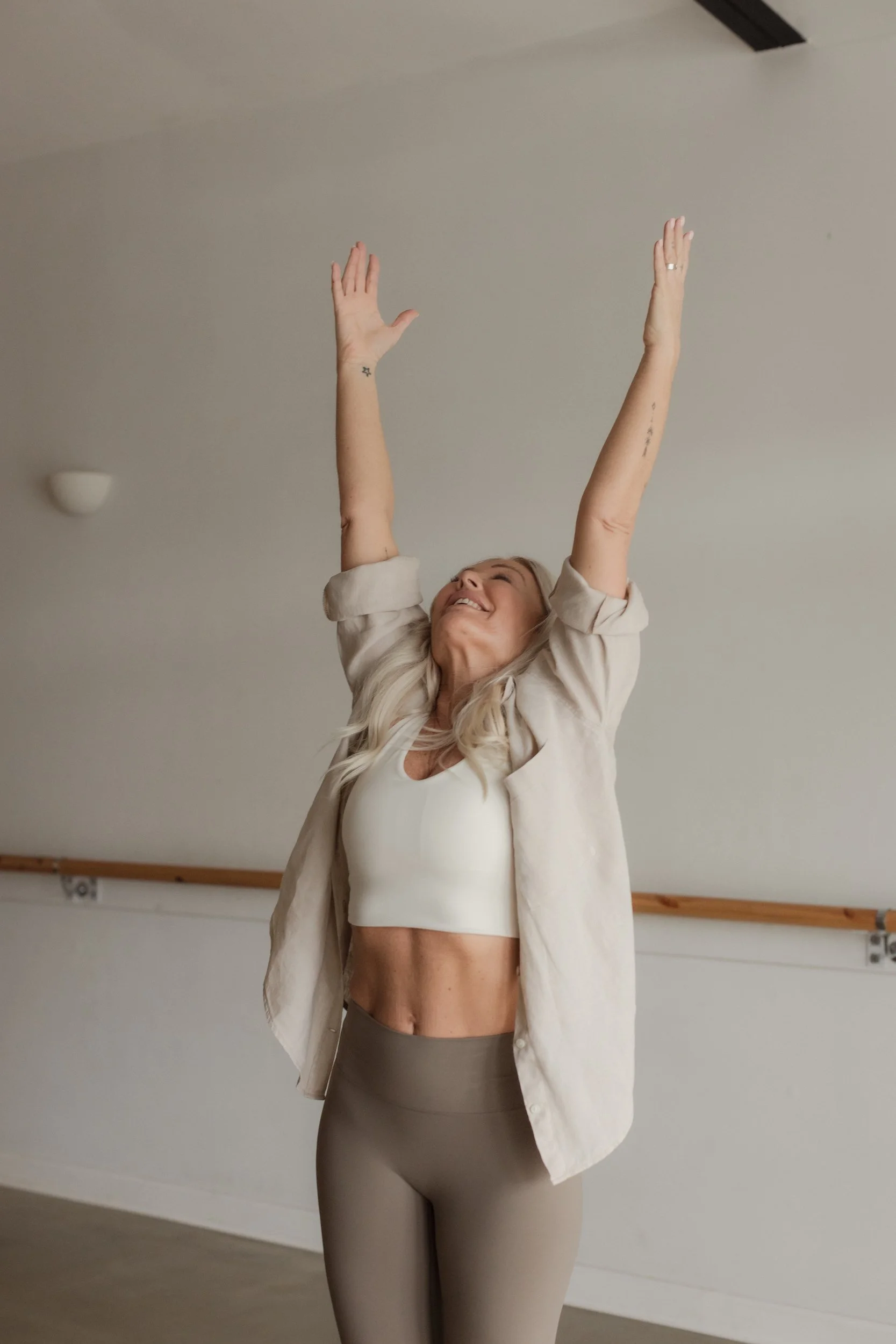 A woman with long blonde hair, wearing a white sports bra, beige leggings, and an open beige shirt, smiling with her arms raised above her head in a dance studio or gym.