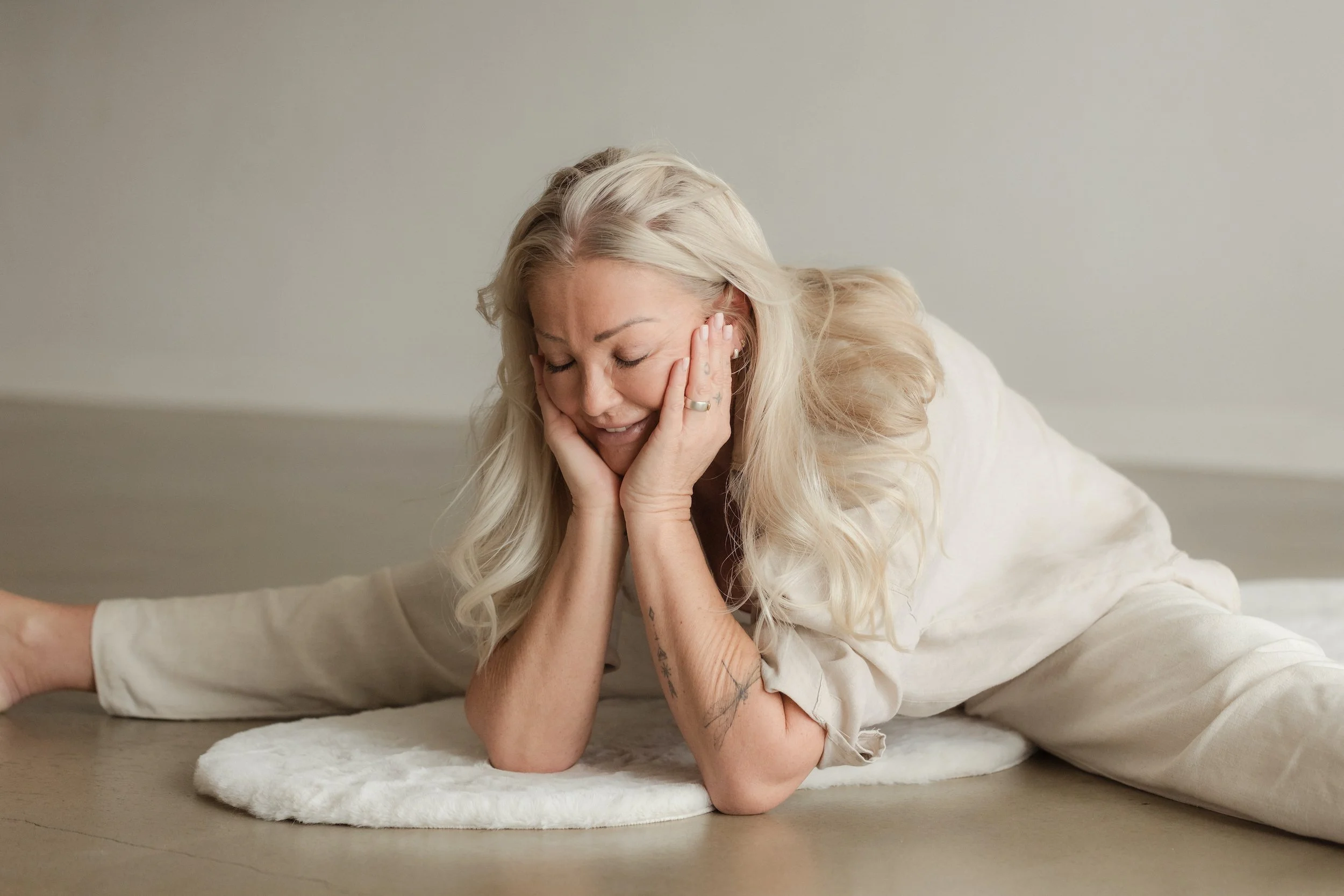 A woman with long blonde hair lying on her side on a white rug, leaning on her left hand, with her eyes closed and a gentle smile.