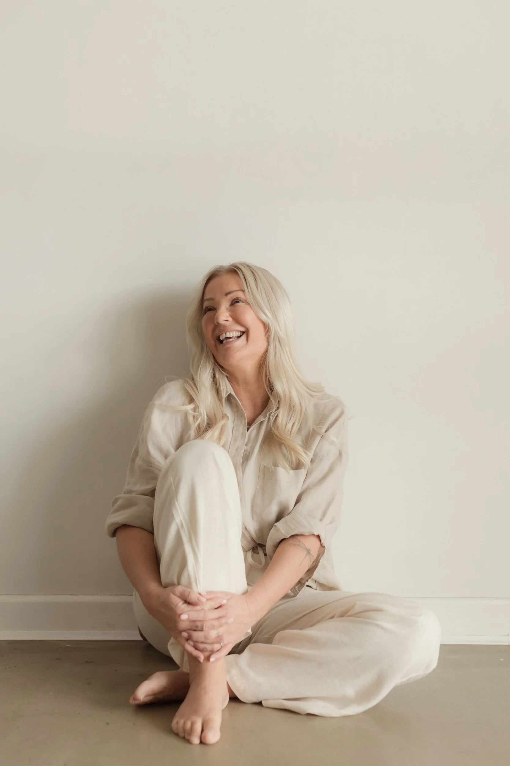 A woman with long blonde hair sitting on the floor against a light-colored wall, laughing, wearing a beige shirt and pants, with her legs crossed and hands clasped around her knee.