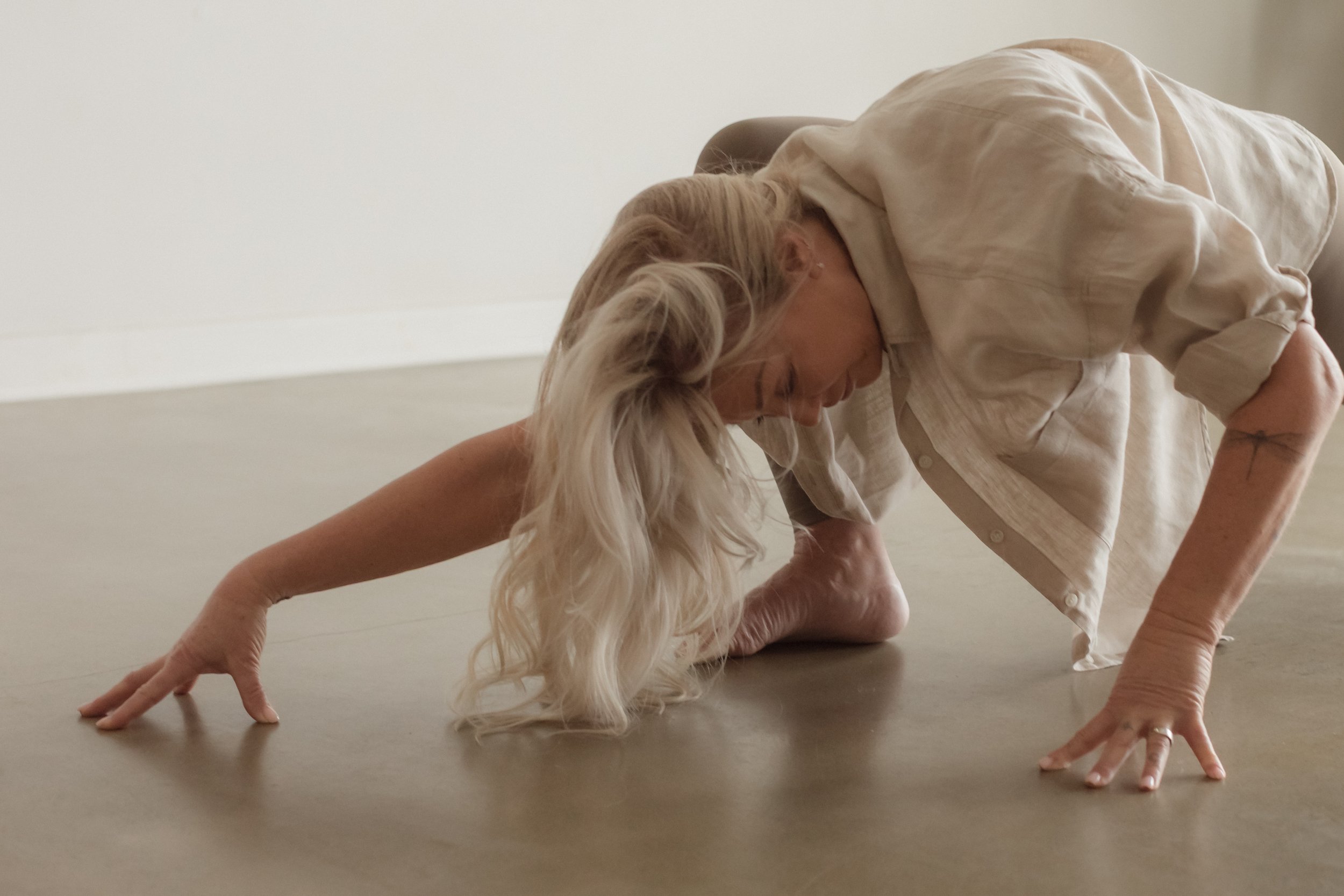 A woman with long blonde hair practicing yoga in a plank position on the floor, wearing a beige shirt, in a minimalist room with light-colored walls and floor.