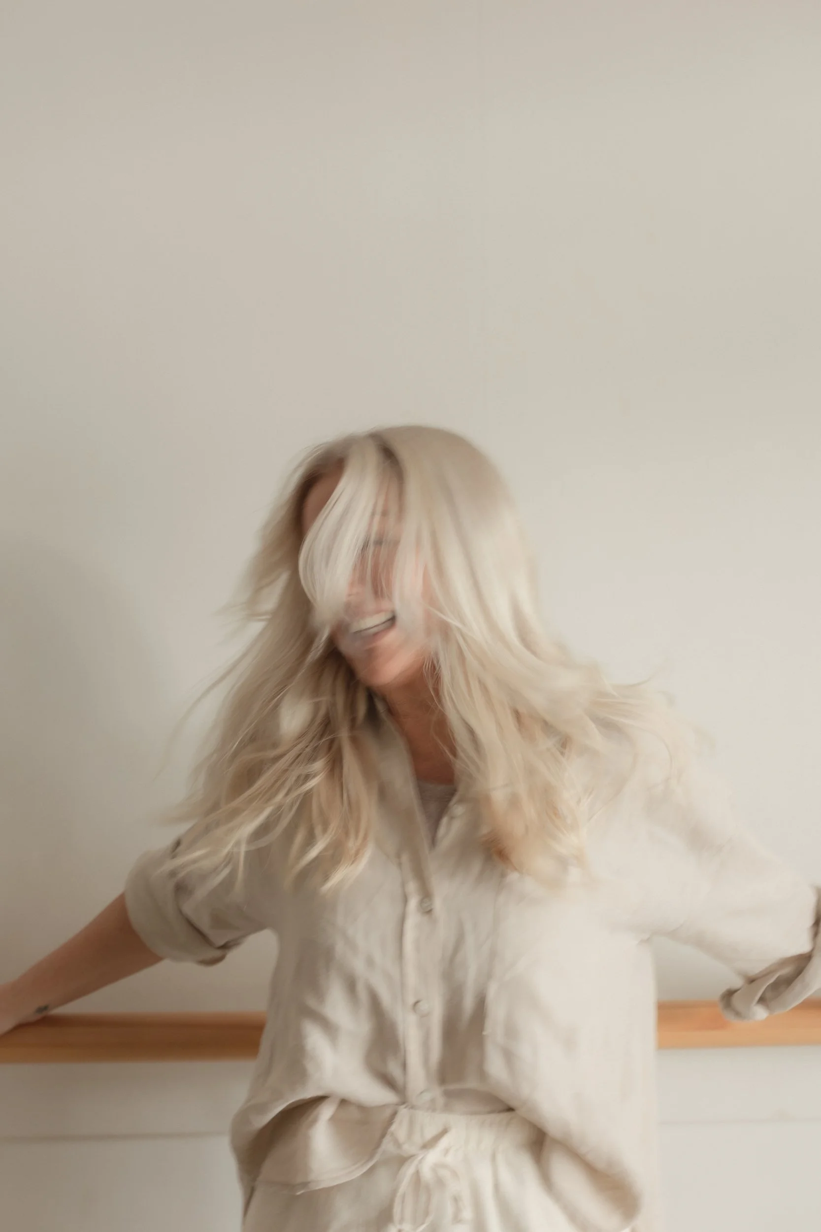 A person with long, wavy blonde hair and glasses, wearing a light-colored shirt, standing in front of a plain, off-white wall.