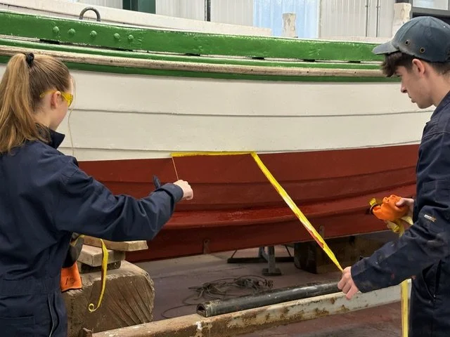 Two people working on a boat in a workshop, measuring with a tape measure.