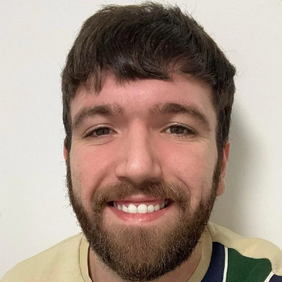A smiling man with a beard and short brown hair standing against a plain white wall.