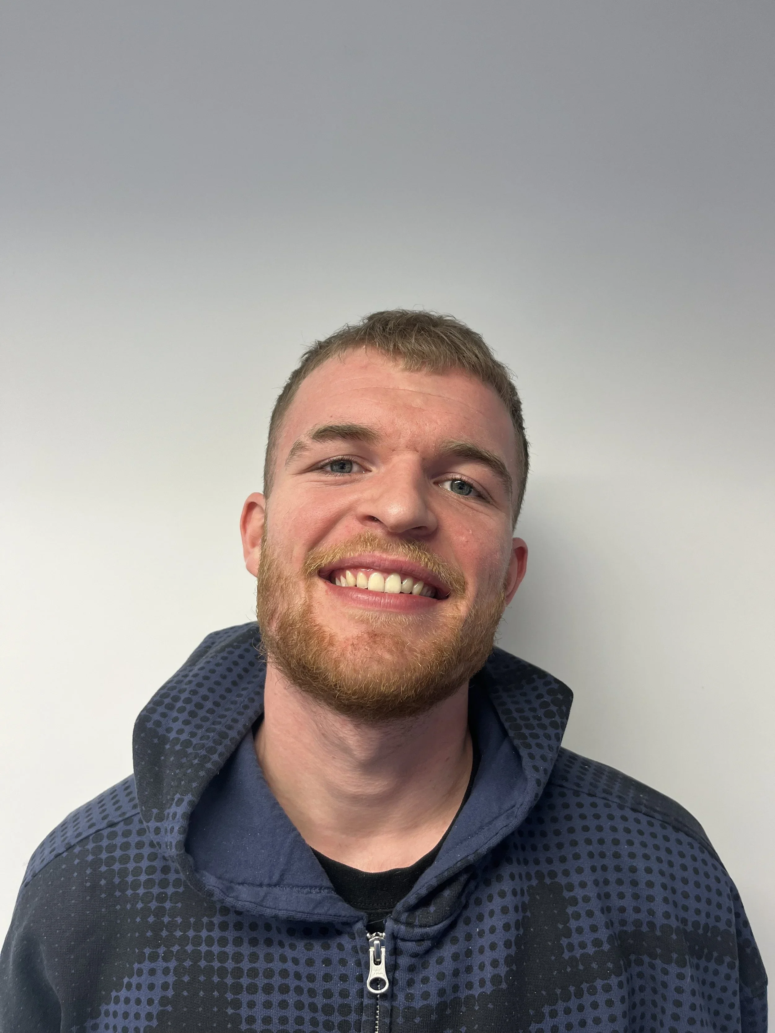 A young man with light skin, short light brown hair, a beard, and blue eyes smiling against a plain white wall. He is wearing a dark blue hoodie with a zipper and patterned details.