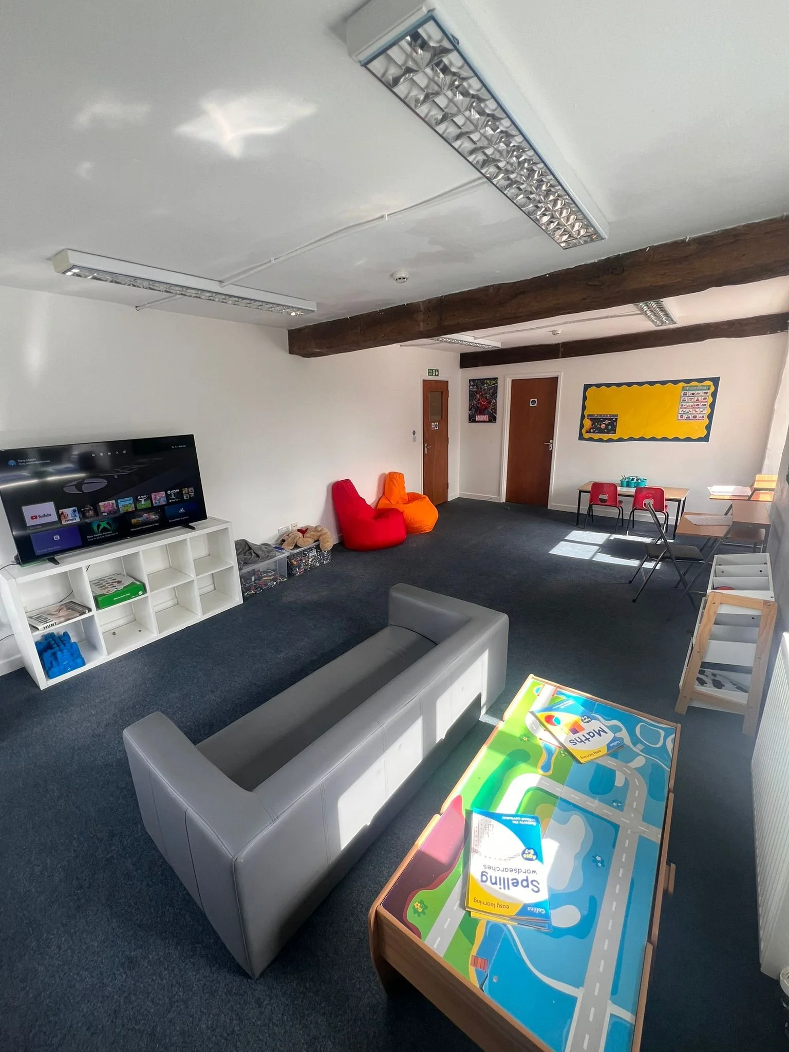 A children’s playroom with a gray couch, a colorful mat with road illustrations, a white cubical shelf with toys, a flat-screen TV, and small chairs at a table with toys, against white walls and dark carpeted floor. There are bean bag chairs, children's books, and framed pictures on the wall.