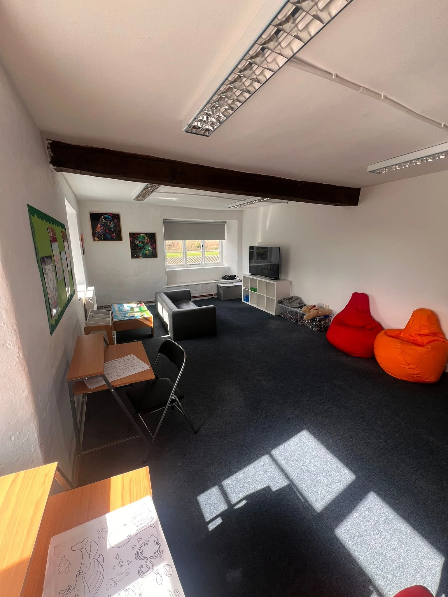 A classroom or activity room with black carpet, white walls, and a window letting in sunlight. There are two colorful bean bags (red and orange) on the right, a small black sofa, a white shelving unit with a TV, and various arts and crafts supplies. A table with chairs is on the left, and some colorful artwork and a green bulletin board are on the walls.